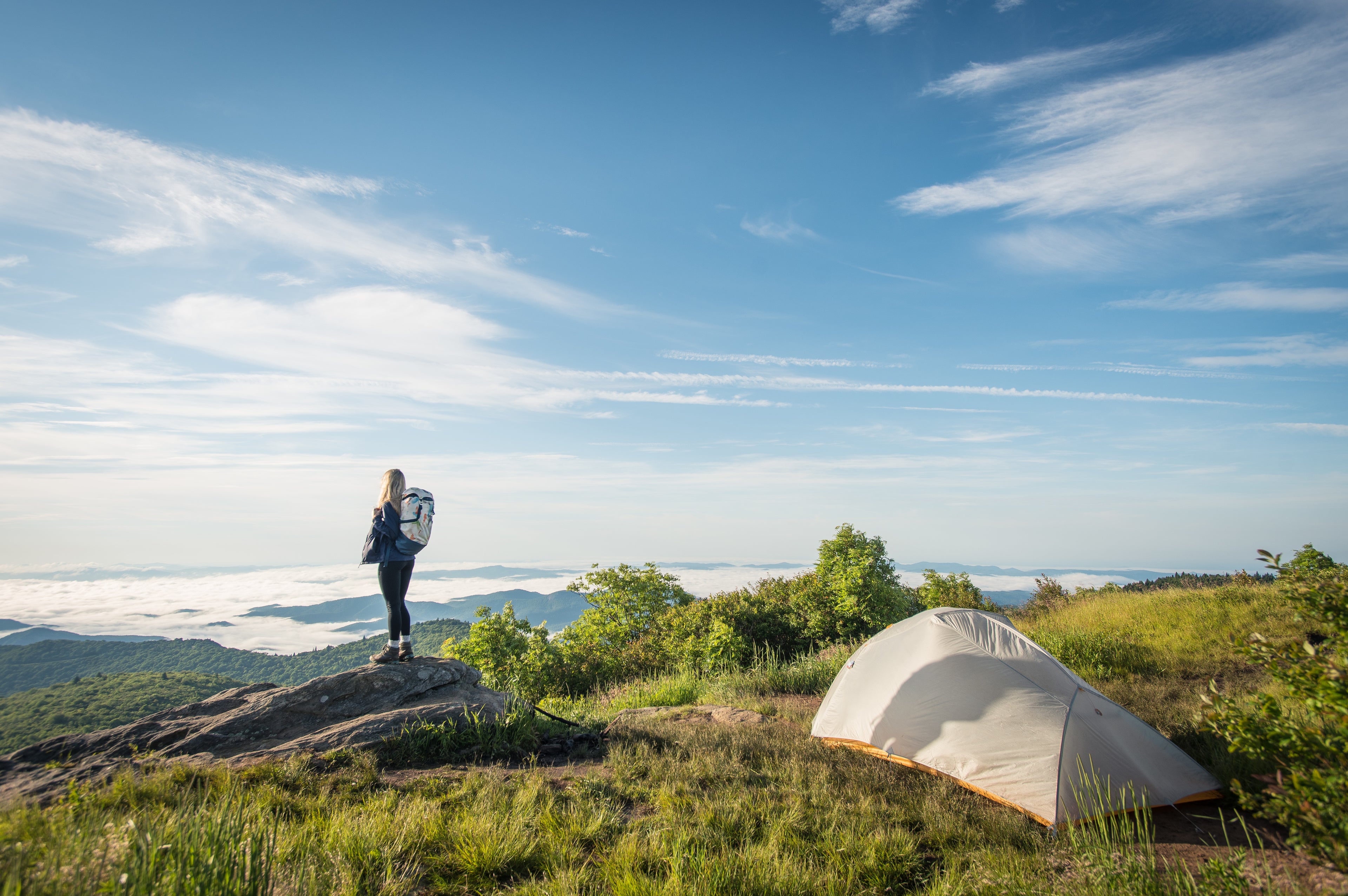 A person with a backpack stands on a rock overlooking a scenic mountain landscape, beside a pitched tent on grassy ground under a bright blue sky with thin clouds.
